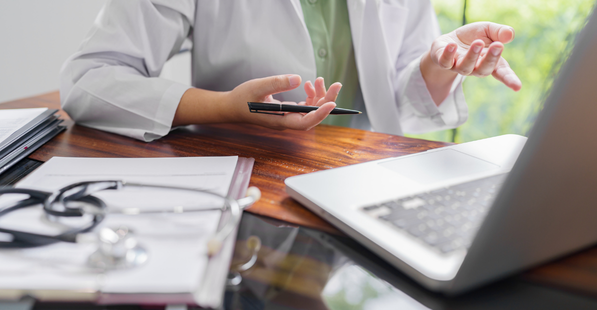 Stock image of a person working at a laptop with a notebook and a stethoscope next to them on the desk